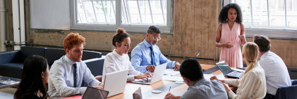 Executives gatherd around the tabe, working at laptops, one women standing and gesturing toward one for comment,